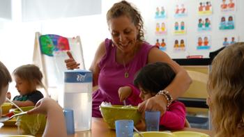 educator smiling and helping a preschooler serve herself fruits